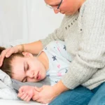 En la imagen se muestra a una mujer cuidando de una chica que parece ser menor de edad. La chica se encuentra acostada de lado en una cama.