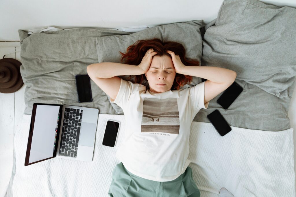 En la fotografía aparece una mujer en plano medio acostada sobre una almohada gris, de cabello castaño oscuro con las manos en la cabeza y ella rodeada de smartphones y un portatil.