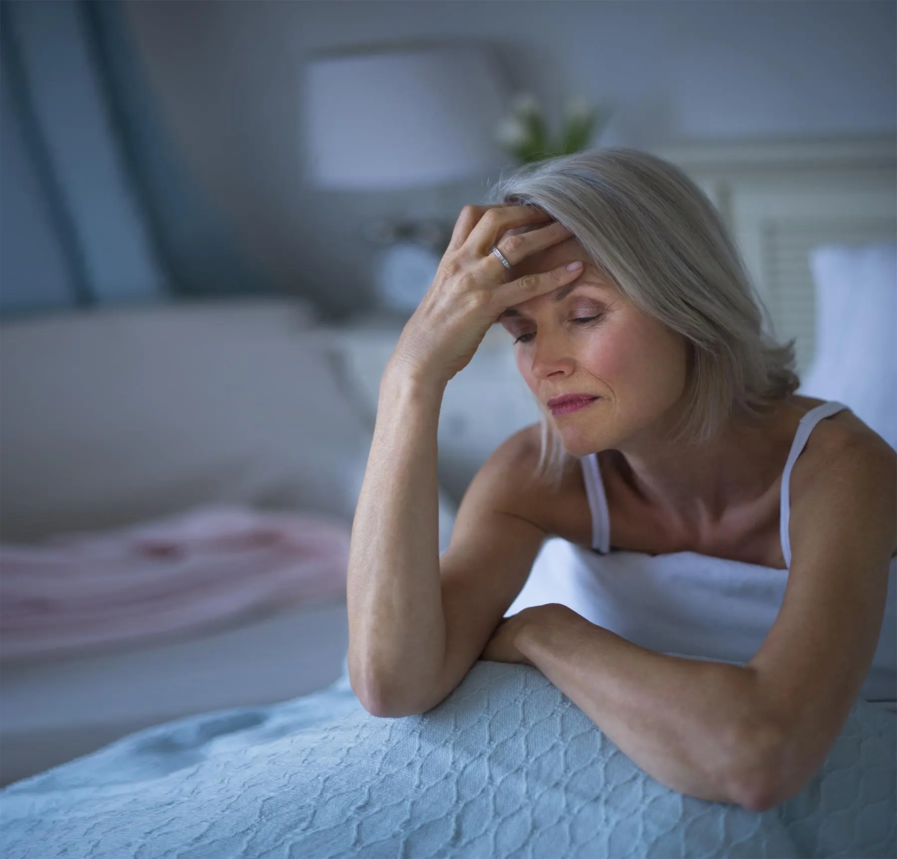 En la imagen se muestra una mujer sentada en su cama algo estresada por no dormir, con la mano sosteniéndole la cabeza y con los ojos cerrados.