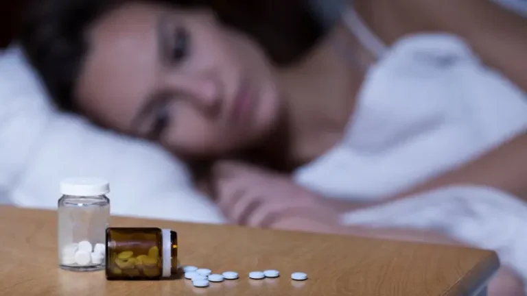 Fotografía de una mujer acostada con los ojos abiertos, acostada en la cama y arropada con sábanas blancas, está mirando hacia abajo, y dos francos de pastas sobre la mesita de noche.