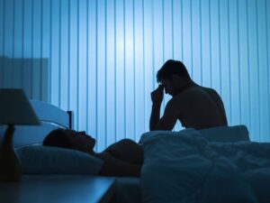 En esta fotografía se muestra a una pareja, mientras la mujer duerme tranquilamente, el hombre está sentado a la orilla de la cama con una mano sobre su cabeza.